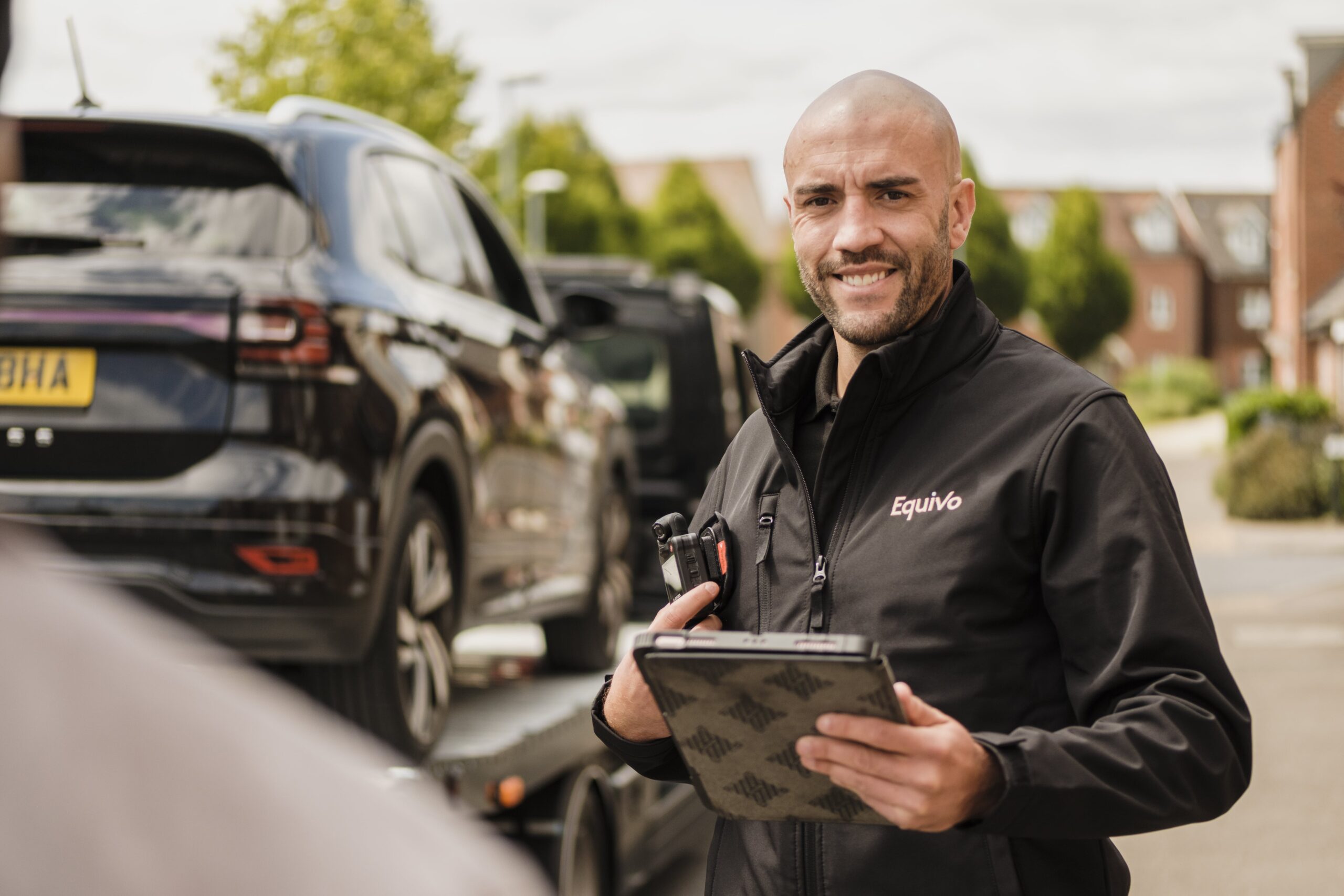 An enforcement agent holds a tablet while standing beside a car designated for repossession.